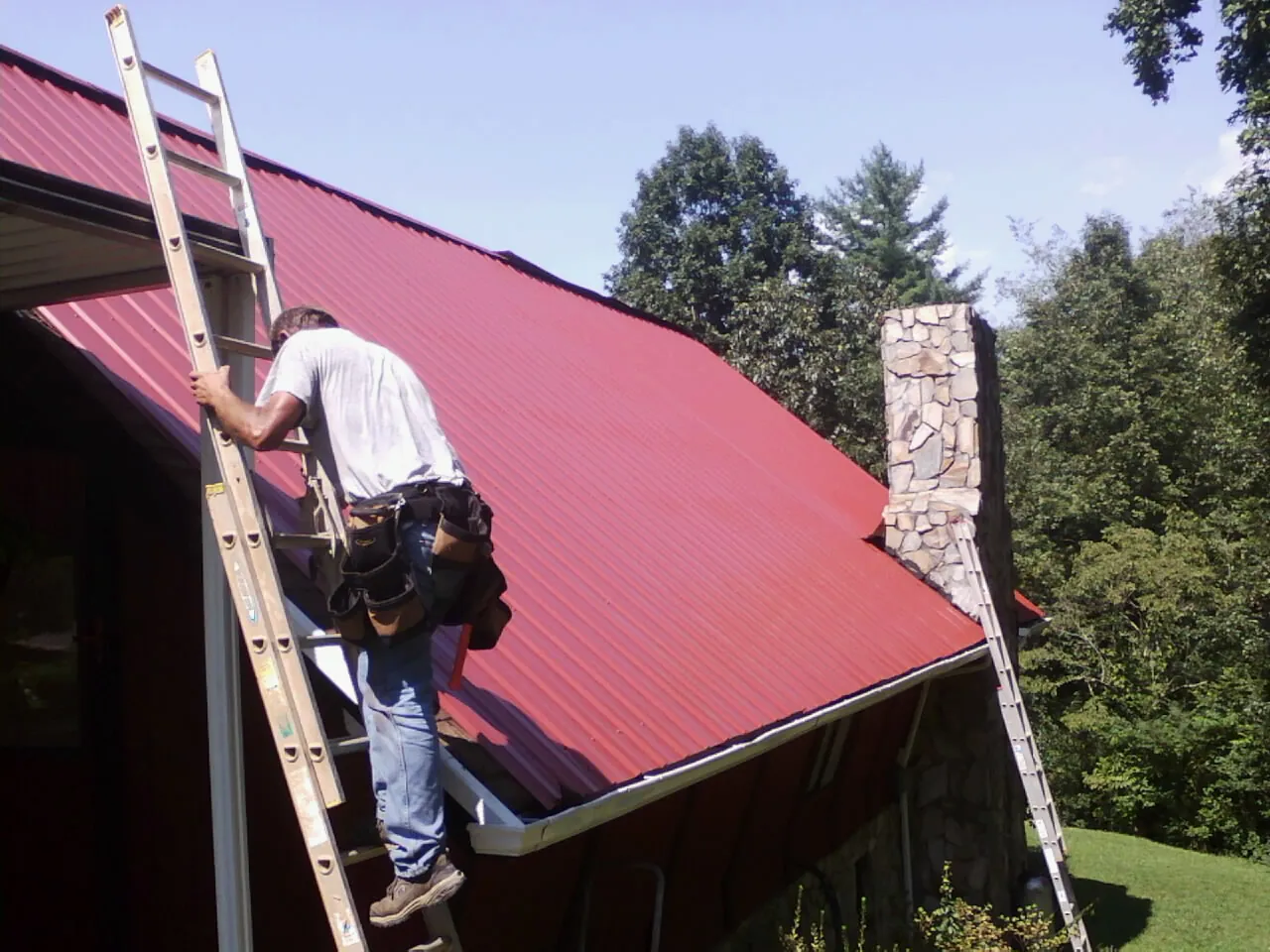 Roofer on a ladder beside a red metal roof and stone chimney — Soffit & Fascia Repair by Roofing Brothers in Factoryville